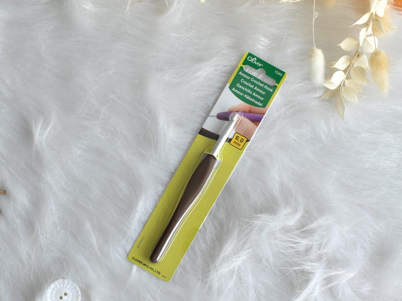 May include: A Clover Amour crochet hook, size 6.0 mm, on a yellow and green card. The hook has a brown handle and a silver metal hook. The packaging includes the text "Amour Crochet Hook" in multiple languages.