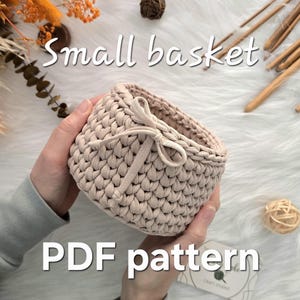 May include: A small, beige crochet basket with a bow detail is held in hands. The basket is made of thick yarn and has a textured appearance. The text "Small basket" and "PDF pattern" are visible in the image.