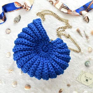 May include: A vibrant blue, crochet seashell-shaped handbag with a gold chain strap. The bag is surrounded by seashells and a blue and white patterned scarf. The bag's design mimics a nautilus shell.
