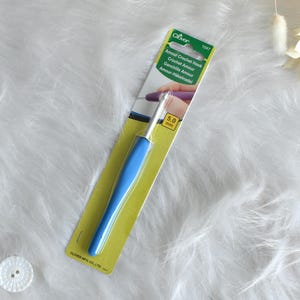 May include: A blue and silver crochet hook on a yellow and green card. The card reads "Amour Crochet Hook" and "5.0 mm". The hook has a blue handle and a silver metal hook. The background is a white, fluffy surface.
