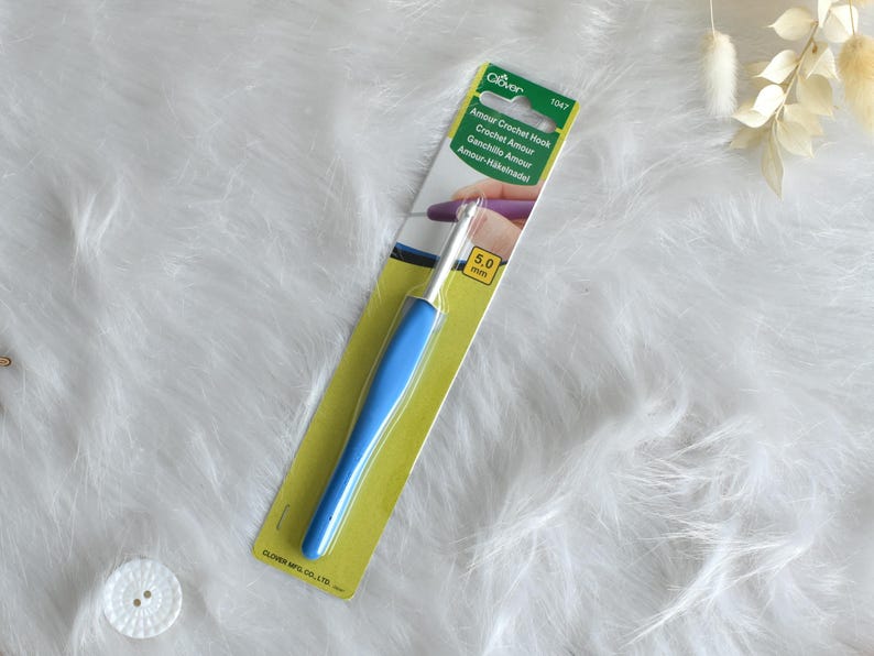 May include: A blue and silver crochet hook on a yellow and green card. The card reads "Amour Crochet Hook" and "5.0 mm". The hook has a blue handle and a silver metal hook. The background is a white, fluffy surface.
