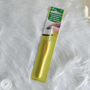 May include: A yellow Clover Amour crochet hook, 2.5 mm, on a green and white card. The hook has a yellow handle and a silver metal hook. The packaging includes the text "Amour Crochet Hook" in multiple languages.