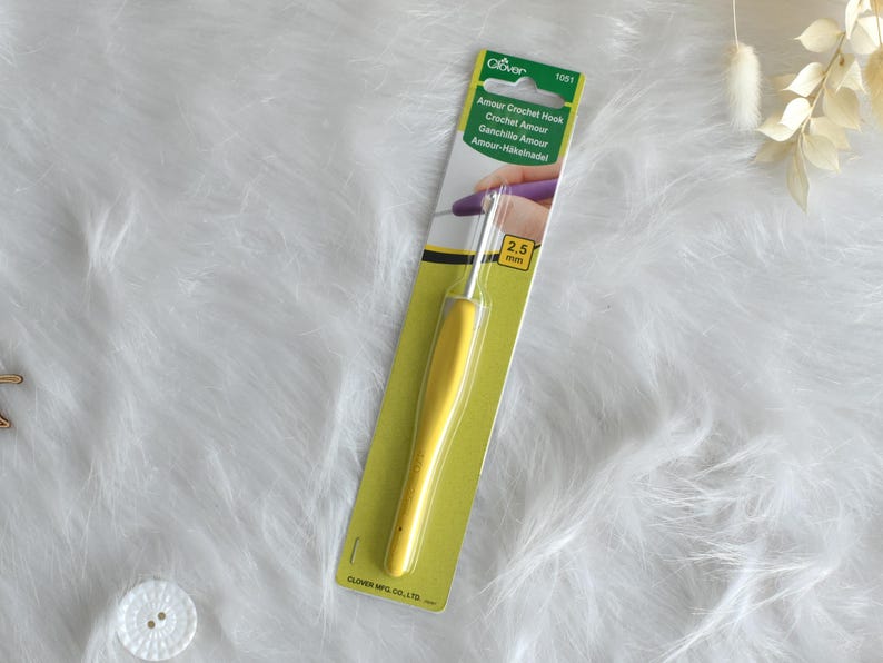 May include: A yellow Clover Amour crochet hook, 2.5 mm, on a green and white card. The hook has a yellow handle and a silver metal hook. The packaging includes the text "Amour Crochet Hook" in multiple languages.