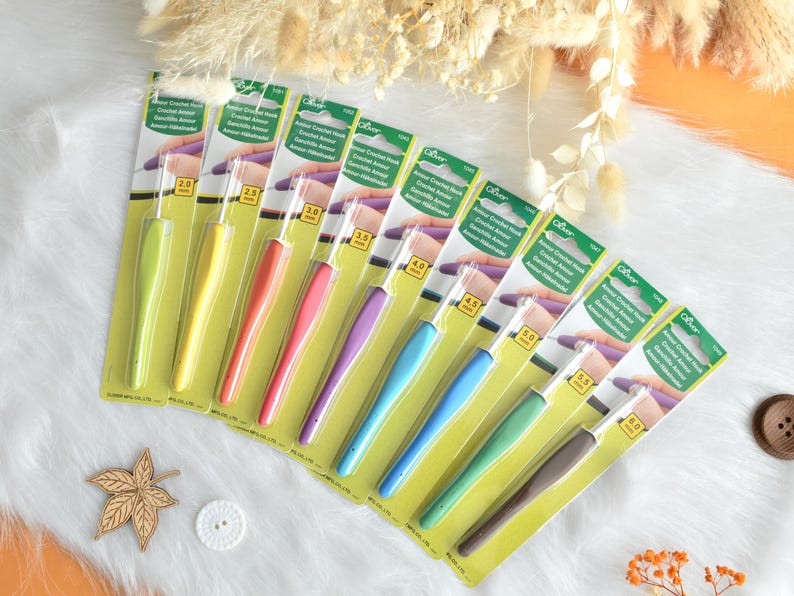 May include: A collection of Clover Amour crochet hooks in various sizes and colors, including green, yellow, red, purple, blue, and brown. Each hook is packaged in a green and white card with the hook size clearly marked.
