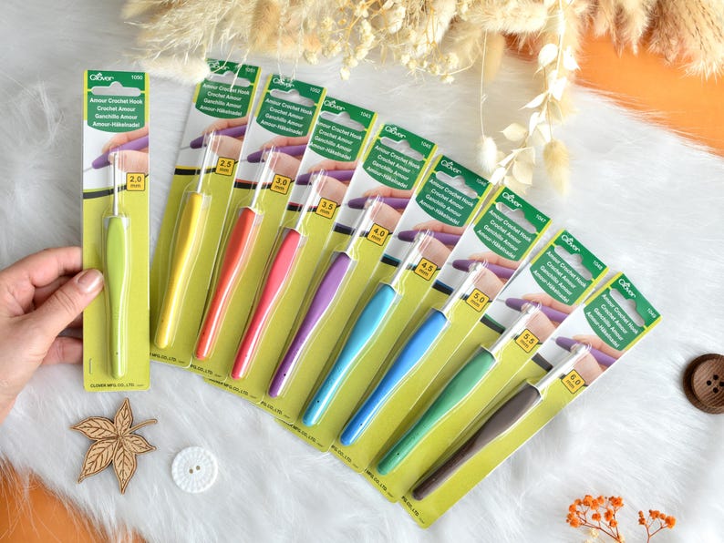 May include: A collection of Clover Amour crochet hooks in various sizes, each with a colorful, ergonomic handle. The hooks range from 2.0 mm to 6.0 mm, displayed on a white surface with decorative elements.