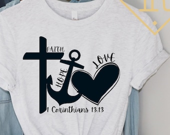 May include: White t-shirt with a black graphic design featuring a cross, an anchor, and a heart. The words "Faith", "Hope", and "Love" are written on the graphic. The text "1 Corinthians 13:13" is also included.