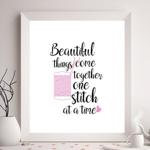 Puede incluir: Una impresión enmarcada blanca con una cita en negro y rosa que dice "Beautiful things come together one stitch at a time". Un corazón rosa está al final de la cita. Un carrete de hilo rosa y una aguja están ilustrados junto a la cita.