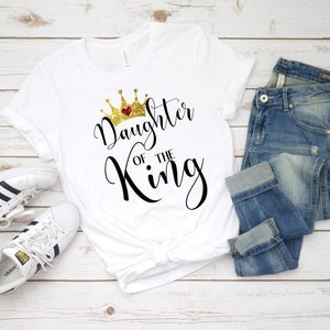 May include: White t-shirt with a gold crown and the text "Daughter of the King" in black script.
