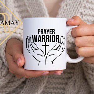 May include: White ceramic mug with a black design of two hands holding a cross with the text "PRAYER WARRIOR" above the cross.