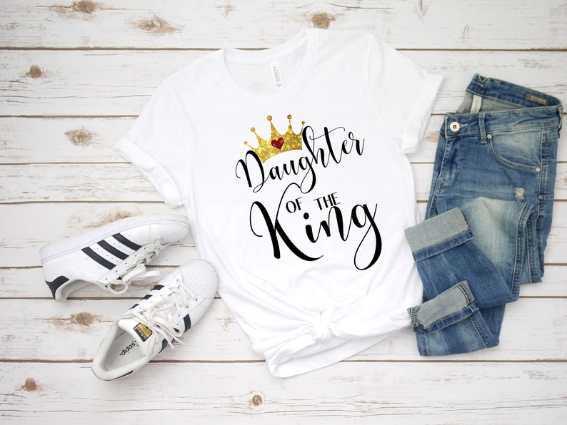 May include: White t-shirt with a gold glitter crown and the text "Daughter of the King" in black script.