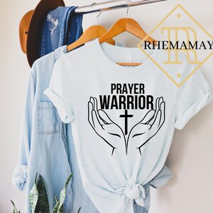 May include: A light blue t-shirt with the text "Prayer Warrior" and a cross in between two hands in a praying position.