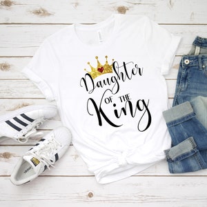 May include: White t-shirt with a gold glitter crown and the text "Daughter of the King" in black script.