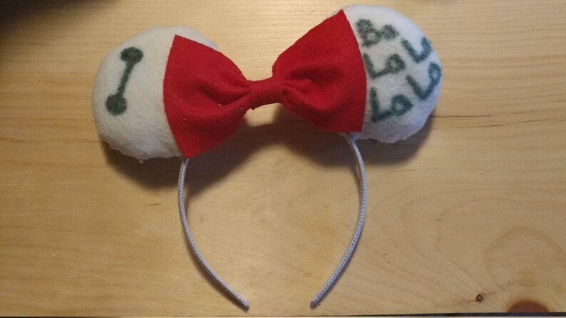 May include: White and red fabric headband with a bow. The headband has a white fabric circle with a green outline of a robot face on one side and a white fabric circle with green text on the other side.