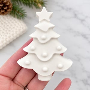 May include: A matte white, three-tiered Christmas tree ornament featuring a star topper and small, raised circular details on each branch level. The tree has a smooth, solid surface and a cylindrical base at the bottom, presented in a flat, stylized form.