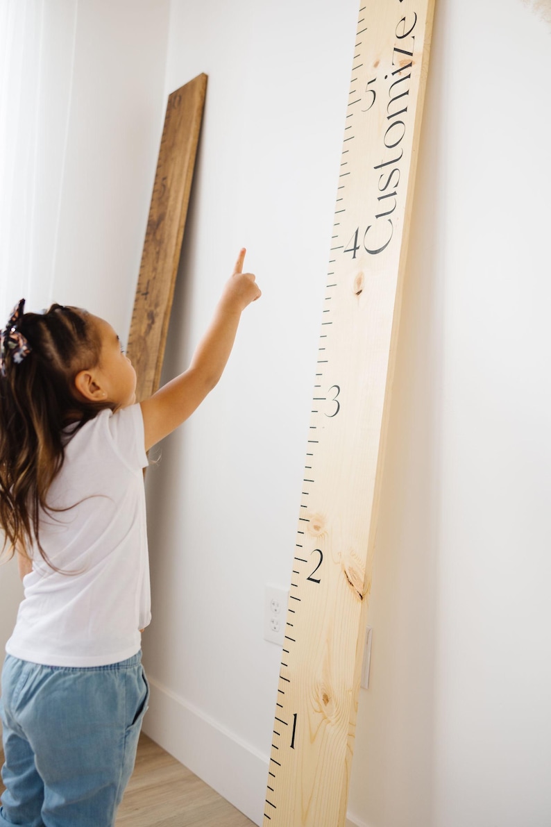 Puede incluir: Un ni&ntilde;o mide su altura en una tabla de crecimiento de madera con marcas negras. La tabla est&aacute; etiquetada como "Personalizar" en la parte superior.