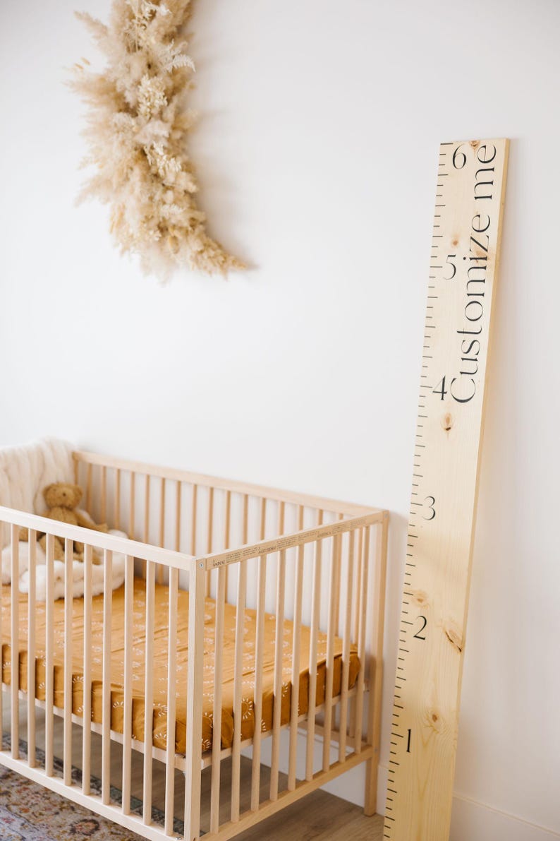 Puede incluir: Un gr&aacute;fico de crecimiento de madera con el texto "Customize me" impreso en &eacute;l. El gr&aacute;fico est&aacute; montado en una pared blanca junto a una cuna con una s&aacute;bana de estampado marr&oacute;n y blanco.