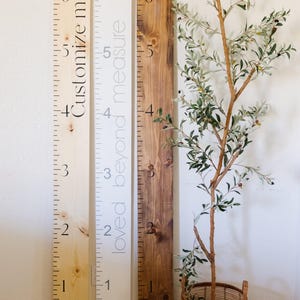 May include: Three wooden growth charts standing vertically. The left board is light wood with the text Customize me. The center board is white with the text loved beyond measure. The right board is dark stained wood. All feature black markings up to 6 feet.