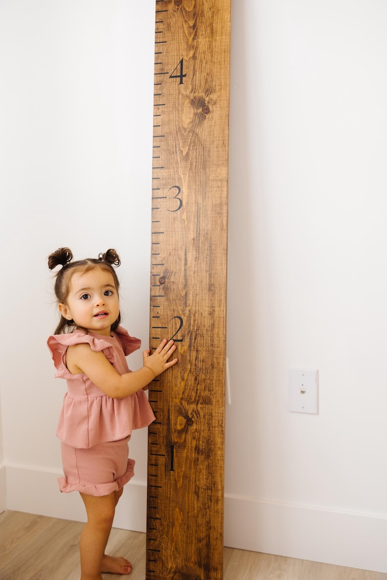 Puede incluir: Una ni&ntilde;a peque&ntilde;a se para junto a una tabla de crecimiento de madera con n&uacute;meros negros. La tabla est&aacute; marcada con los n&uacute;meros del 1 al 4. La ni&ntilde;a lleva un atuendo rosa con volantes y tiene el pelo recogido en dos coletas.