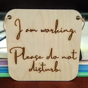 May include: A wooden sign with the text "I am working. Please do not disturb." The sign is light brown and has a string attached to the top.