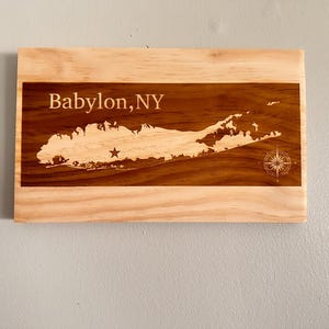 May include: Wooden plaque featuring a map of Long Island, New York, with the text "Babylon, NY" engraved at the top. The map is detailed with a star and compass rose design. The plaque is rectangular with a light wood border.