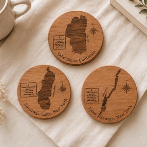 May include: Three circular wooden coasters featuring laser-engraved maps of Lake Tahoe, Saratoga Lake, and Lake George. Each coaster displays the lake name, location, coordinates, depth, and a compass rose. The wood grain is visible throughout.