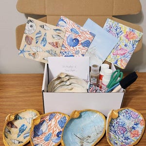 Peut inclure: Un kit de bricolage avec des coquillages peints, de la peinture, des pinceaux et du papier à motifs. Les coquillages sont décorés de divers motifs et couleurs, dont le bleu, l'orange et l'or. Le kit est dans une boîte blanche à l'intérieur d'une boîte en carton.