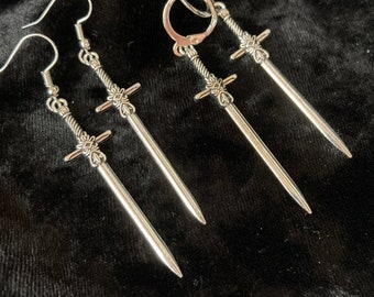 May include: A pair of silver sword earrings with a hoop style back. The earrings are on a black velvet background.