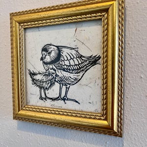 May include: A framed art print showing a black and white linocut of two birds. The larger bird shelters the smaller one. The gold frame has a textured, rope-like pattern around the edges.