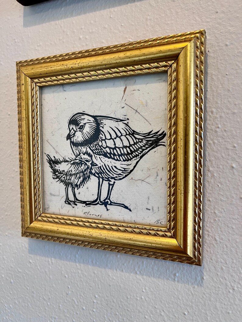 May include: A framed art print showing a black and white linocut of two birds. The larger bird shelters the smaller one. The gold frame has a textured, rope-like pattern around the edges.