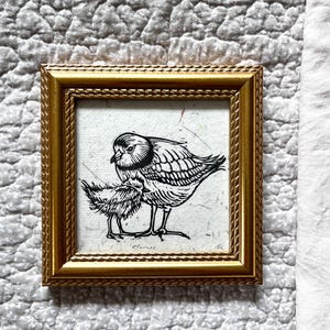 May include: A square framed art print featuring a black ink linocut illustration of an adult bird and a small chick on textured off-white paper. The frame is metallic gold with a rope-like embossed border. The word Plovers is written in pencil.