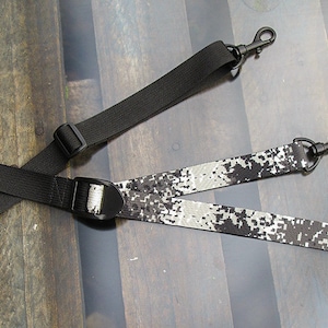 Könnte beinhalten: Ein schwarzer und weißer Kameragurt mit schwarzen verstellbaren Schnallen und Metallclips.