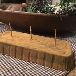 May include: A rustic, rectangular, brown and green metal candle holder with three wooden candle holders. The candle holder is sitting on a checkered tablecloth.
