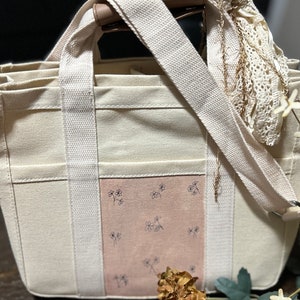 May include: A white canvas tote bag with a pink floral design and a long strap. The bag has multiple pockets and a zipper closure.