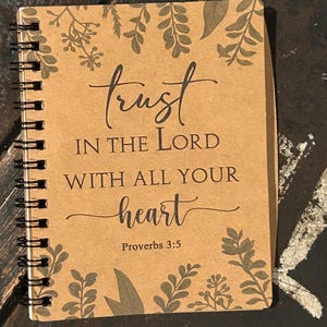 Może przedstawiać: Spiralny notatnik z okładką z papieru typu kraft. Okładka zawiera słowa "trust IN THE LORD WITH ALL YOUR heart" i "Proverbs 3:5" w czarnym skrypcie. Ozdobne gałązki liści otaczają tekst.
