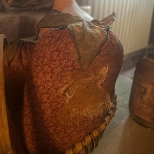 Puede incluir: Una cesta rústica contiene dos calabazas de tela. Las calabazas son de color rojo oscuro con un estampado floral y un parche de arpillera. Los tallos están cubiertos con una tela a cuadros. La cesta es de madera tejida.