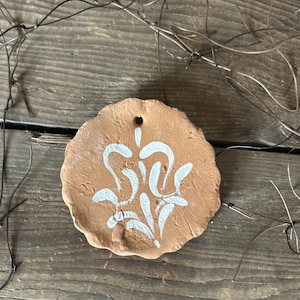 Könnte beinhalten: Ein Terrakotta-Anhänger mit einem weißen Blumenmuster. Der Anhänger hat oben ein Loch zum Aufhängen.