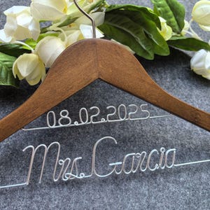 May include: A wooden wedding dress hanger with the date 08.02.2025 and the name Mrs. Garcia in silver wire. The hanger is decorated with white flowers and green leaves.