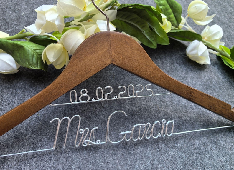 May include: A wooden wedding dress hanger with the date 08.02.2025 and the name Mrs. Garcia in silver wire. The hanger is decorated with white flowers and green leaves.