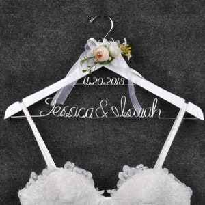 May include: White wooden wedding dress hanger with the names "Jessica & Isaiah" in wire script and the date "11.20.2018". The hanger is decorated with a floral arrangement and a sheer ribbon. A white lace wedding dress is partially visible.