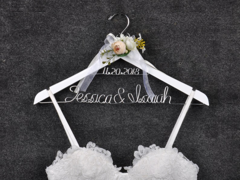 May include: White wooden wedding dress hanger with the names "Jessica & Isaiah" in wire script and the date "11.20.2018". The hanger is decorated with a floral arrangement and a sheer ribbon. A white lace wedding dress is partially visible.