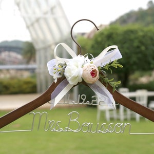 May include: A wooden wedding dress hanger with a metal hook and wire name. The hanger is decorated with white and peach-colored flowers, greenery, and a sheer ribbon. The date 11.19.22 is displayed above the name "Mrs. Bousson".