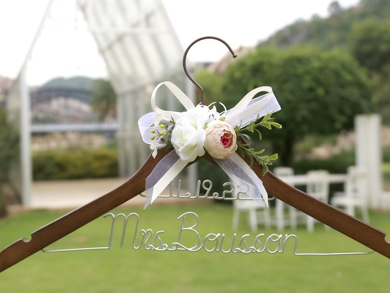 May include: A wooden wedding dress hanger with a metal hook and wire name. The hanger is decorated with white and peach-colored flowers, greenery, and a sheer ribbon. The date 11.19.22 is displayed above the name "Mrs. Bousson".