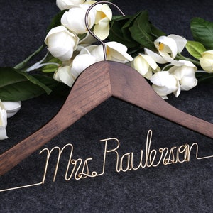 May include: A wooden hanger with a gold wire inscription that reads "Mrs. Rawlerson". The hanger is decorated with white flowers.