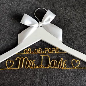 May include: A white wooden hanger with a silver hook and a white satin bow. The hanger features gold wire script that reads "06.06.2026 Mrs. Davis" with heart accents, perfect for a wedding.