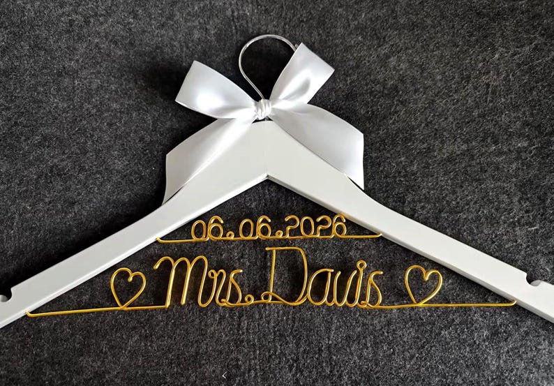 May include: A white wooden hanger with a silver hook and a white satin bow. The hanger features gold wire script that reads "06.06.2026 Mrs. Davis" with heart accents, perfect for a wedding.