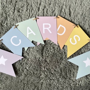 K&ouml;nnte beinhalten: Ein pastellfarbenes Banner mit dem Wort "CARDS" auf einzelnen Flaggen. Die Flaggen sind in Blaut&ouml;nen, Gr&uuml;n, Gelb und Rosa.