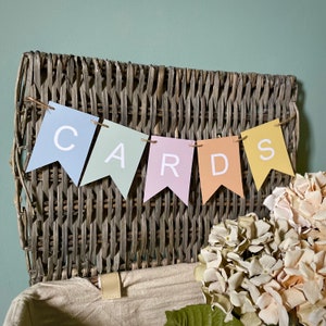 K&ouml;nnte beinhalten: Ein Weidenkorb mit einem Banner, auf dem "CARDS" in bunten Buchstaben steht. Das Banner ist aus Papier und mit Schnur befestigt. Der Korb steht auf einem Tisch mit einem Strau&szlig; wei&szlig;er Blumen im Vordergrund.