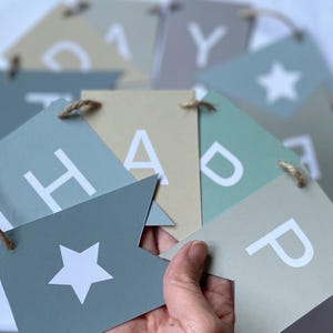 Puede incluir: Una pancarta azul claro y beige con las palabras "Happy" y una estrella impresa en ella. La pancarta est&aacute; atada con un cordel.