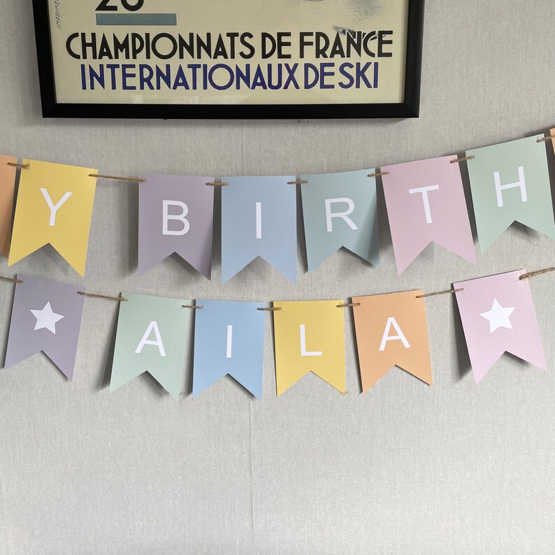 Op de afbeelding: Een pastelkleurig banner met de woorden "Happy Birthday Aila" en twee witte sterren. De banner is aan een touwtje geregen.