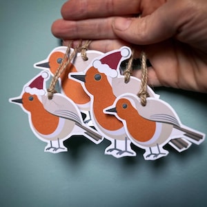 Puede incluir: Una mano sostiene varios adornos navideños. Los adornos tienen forma de petirrojos con pechos naranjas, alas grises y pequeños gorros de Papá Noel. Cada adorno está sujeto a una cuerda de hilo marrón.